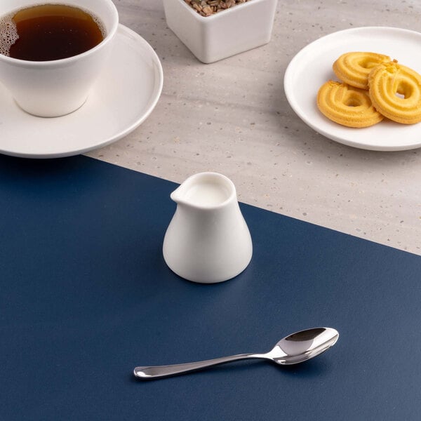 A small white porcelain creamer is placed on a blue and gray surface, surrounded by a cup of tea, a plate of cookies, and a spoon.