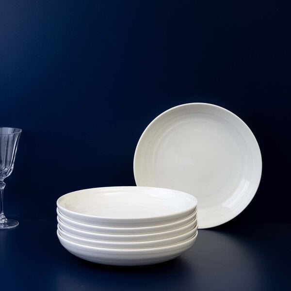 A stack of six round, white shallow bowls is displayed on a dark surface with one bowl standing upright behind the stack and a clear wine glass to the left.