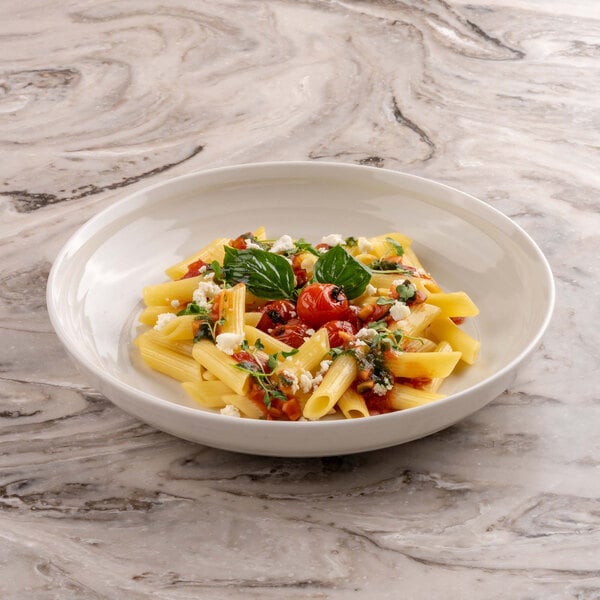 A shallow round ivory bowl filled with penne pasta, cherry tomatoes, crumbled cheese, and fresh basil, placed on a marble surface.