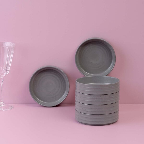 A stack of round gray bowls is displayed on a pink surface, with two additional bowls propped up behind and a clear glass on the left side.
