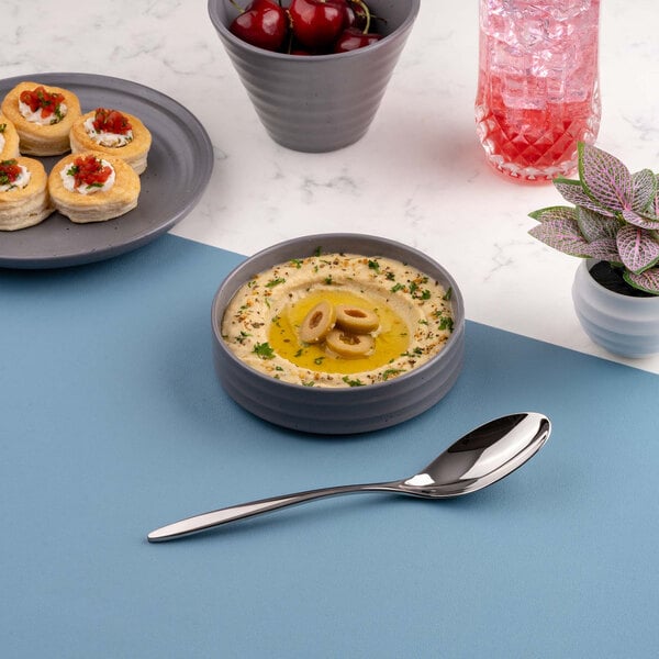 A round gray bowl filled with hummus garnished with olive slices and herbs, placed on a blue surface next to a spoon, with other food items and a plant visible in the background.