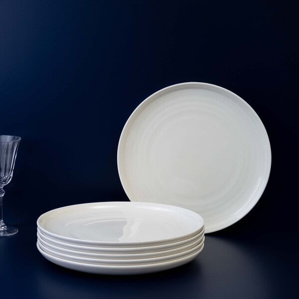 A stack of six round, ivory-colored porcelain plates is shown, with one plate standing upright behind the stack and a clear glass partially visible on the left side.
