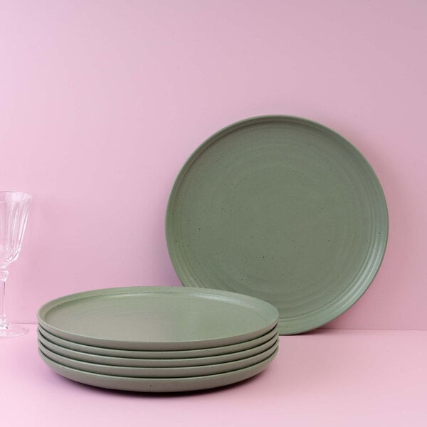 A stack of six round, matte green plates is displayed in front of a pink background, with one plate standing upright behind the stack and a clear glass partially visible on the left.