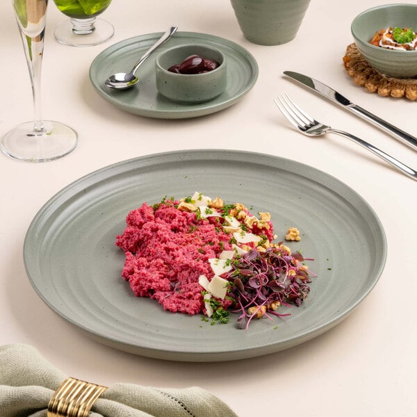 A round, green porcelain plate with a textured surface, featuring a colorful dish with pink, white, and green ingredients, garnished with herbs and nuts.