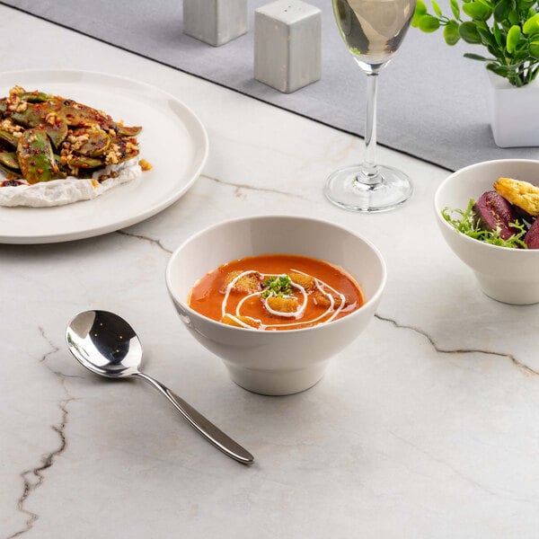 A round ivory bowl filled with tomato soup garnished with cream and herbs, placed on a marble surface alongside a spoon, a plate of food, a smaller bowl with salad, and a glass of white wine.