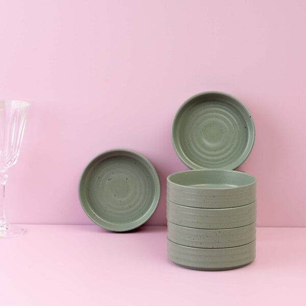 A stack of small, round, green ceramic bowls is displayed against a pink background, with two bowls propped up to show their interiors and a clear glass on the left side.