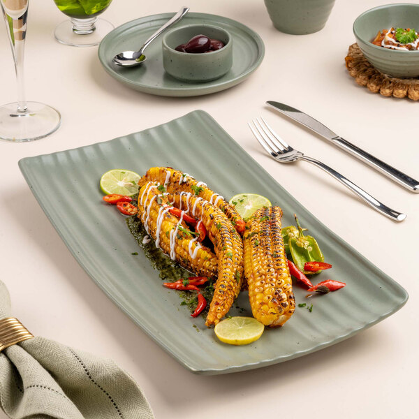 A rectangular green platter with grilled corn on the cob, lime slices, and red chili peppers, set on a table with cutlery and other dishes.