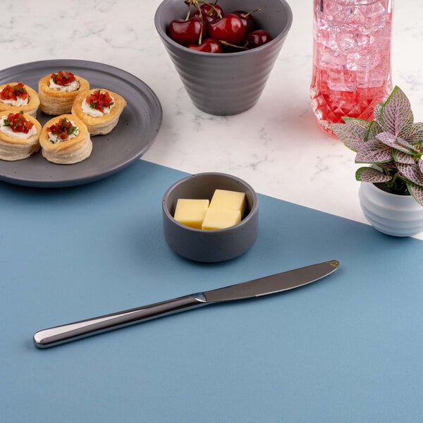 A small round gray bowl containing three pieces of butter is placed on a blue surface, surrounded by a knife, a plate of appetizers, a bowl of cherries, a pink drink, and a potted plant.