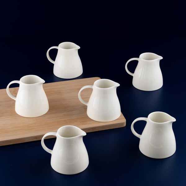 Six small white porcelain creamers are arranged around a wooden board on a dark surface.