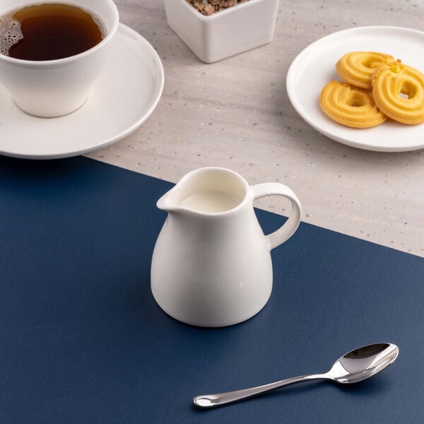 A small white porcelain creamer filled with milk is placed on a blue surface, with a spoon, a cup of tea, and a plate of cookies nearby.