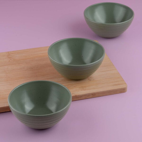 Three green ceramic bowls are arranged on and around a wooden cutting board against a pink background.
