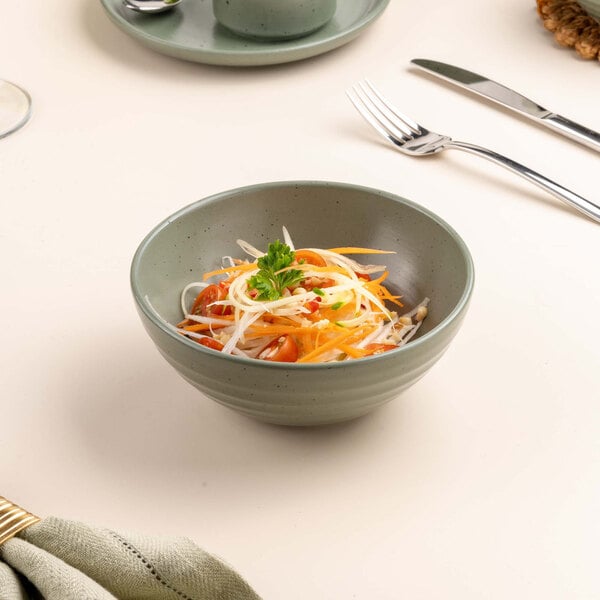 A green porcelain bowl filled with a salad of shredded vegetables and garnished with parsley, placed on a light-colored table with cutlery and a napkin nearby.