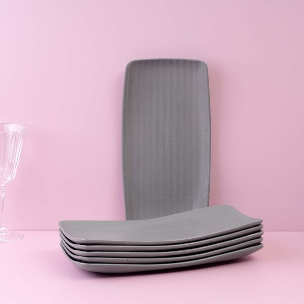 A stack of six rectangular gray plates is displayed in front of a pink background, with one plate standing upright behind the stack and a clear glass on the left side.