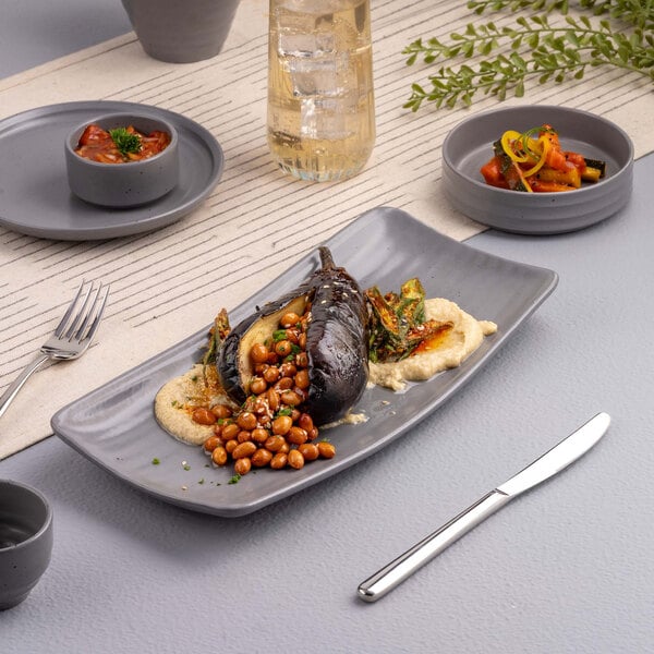 A rectangular gray platter with a serving of roasted eggplant, chickpeas, and a creamy spread, accompanied by side dishes and a drink on a modern table setting.