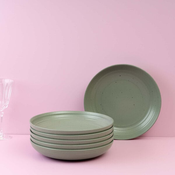 A stack of five light green shallow bowls with a matching bowl standing upright behind them, set against a pink background with a clear glass on the left.