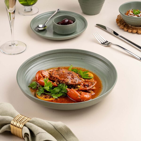 A round, shallow green bowl filled with a crab dish in sauce, garnished with fresh herbs, is placed on a table set with cutlery, a napkin, and other dishes.