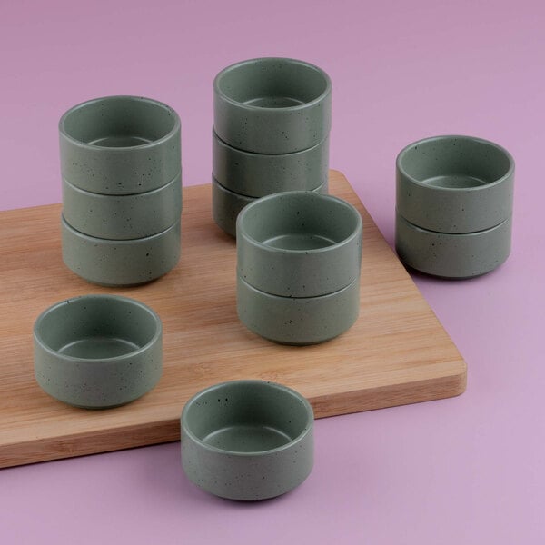 Several small, round, green ceramic bowls are arranged in stacks on and around a wooden board against a pink background.