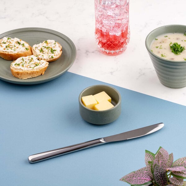 A small green ramekin containing cubes of butter is placed on a blue surface, surrounded by a butter knife, a plate with bread, a bowl of soup, a pink drink, and a plant.