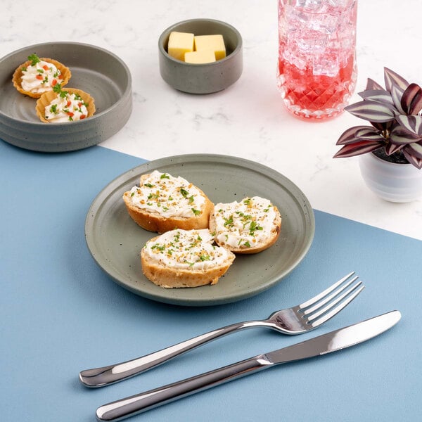 A green round plate with three pieces of bread topped with a creamy spread and herbs, placed on a blue placemat with a fork and knife beside it.