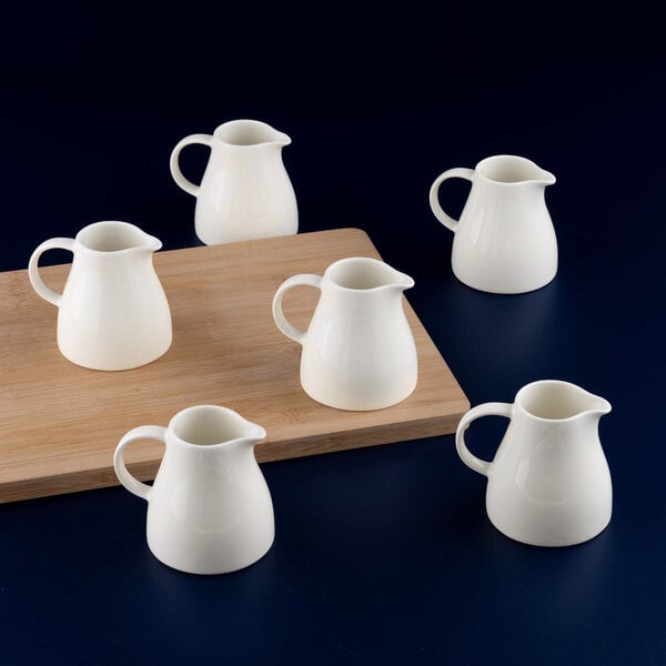 Six small white porcelain creamers are arranged on and around a wooden board against a dark background.