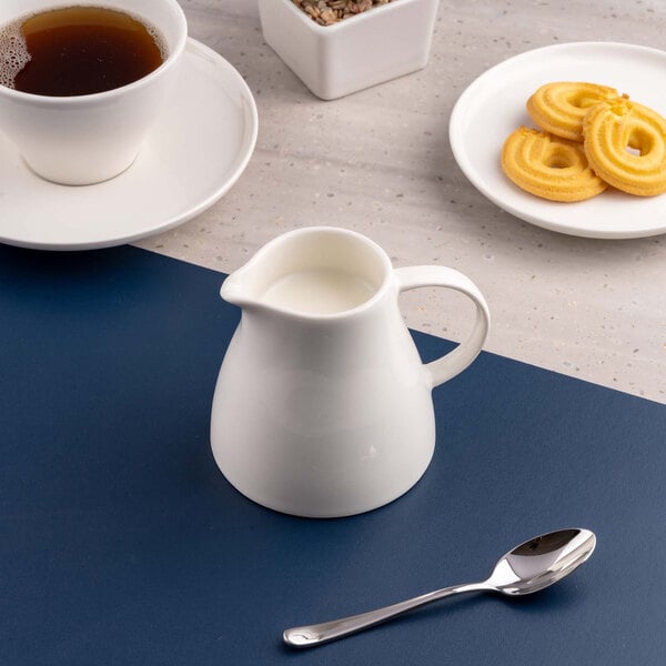 A small white porcelain creamer filled with milk is placed on a blue surface, surrounded by a cup of tea, a plate of cookies, and a spoon.