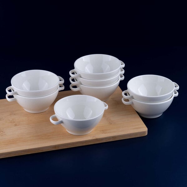 Eight white porcelain soup bowls with handles are stacked in groups on a wooden board against a dark background.