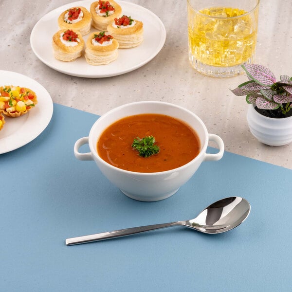 A white porcelain soup bowl with handles filled with tomato soup, garnished with parsley, placed on a blue mat with a spoon beside it; other food items, a drink, and a small potted plant are also visible.
