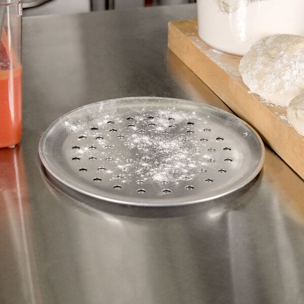 An American Metalcraft aluminum pizza pan with holes in it next to dough and a cutting board.