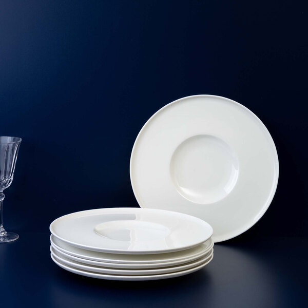 A stack of five white round plates with a single matching plate standing upright behind them, set against a dark blue background with a clear wine glass partially visible on the left.