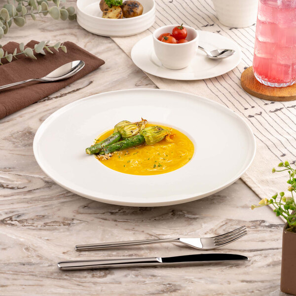 A large white round plate with a gourmet dish featuring yellow sauce and stuffed zucchini flowers, set on a marble table with cutlery, a napkin, and other small dishes nearby.