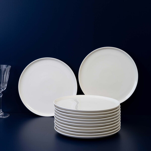 A stack of plain white round plates is displayed on a dark surface with two plates standing upright in the background and a glass on the left side.