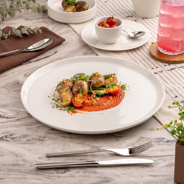 A white round plate with a gourmet dish featuring roasted vegetables and a red sauce, set on a marble table with cutlery and a glass of pink beverage nearby.