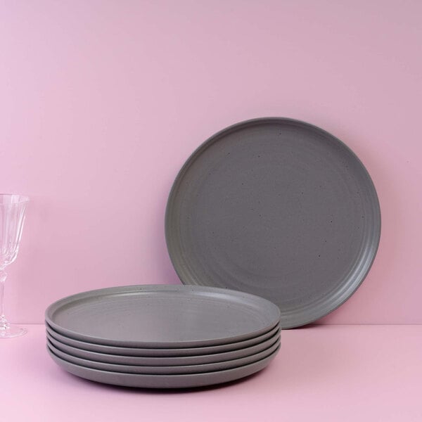 A stack of six round gray plates is displayed in front of a pink background, with one plate standing upright behind the stack and a clear glass visible on the left side.