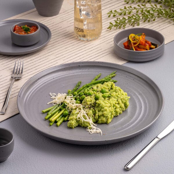 A gray round plate with a serving of green risotto, asparagus, peas, and shredded cheese, set on a modern table with side dishes and a drink.
