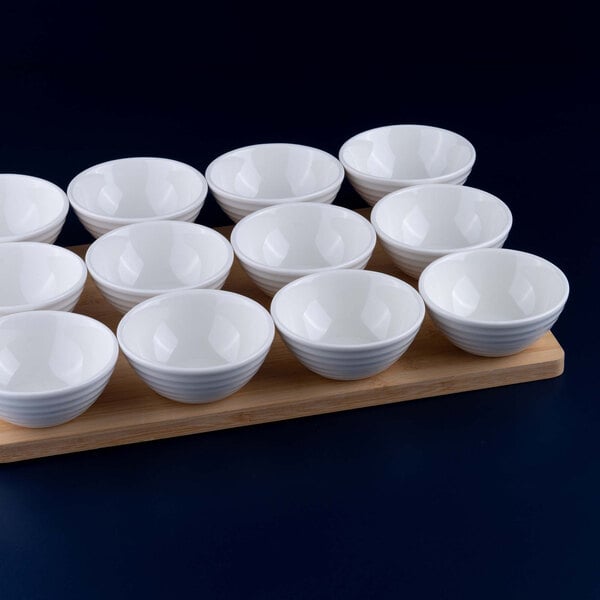 Twelve small white porcelain bowls are arranged on a wooden board against a dark background.