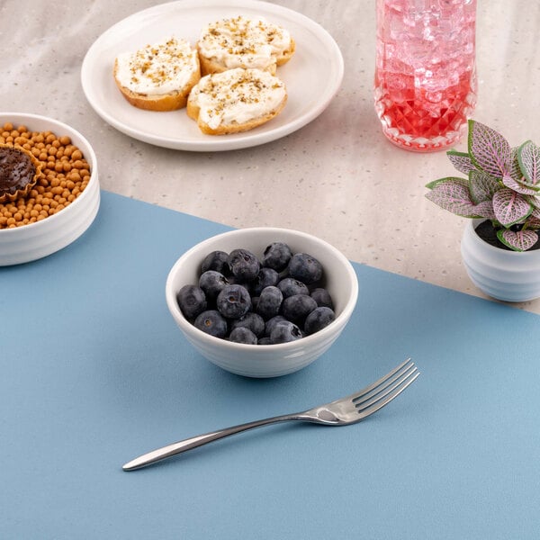 A small white bowl filled with blueberries is placed on a blue surface, surrounded by a fork, a plate with snacks, a glass with a pink drink, another bowl with cereal, and a small potted plant.