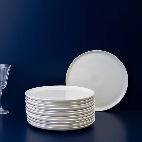 A stack of twelve round, white plates is shown next to a single upright plate and a clear glass on a dark surface with a dark blue background.