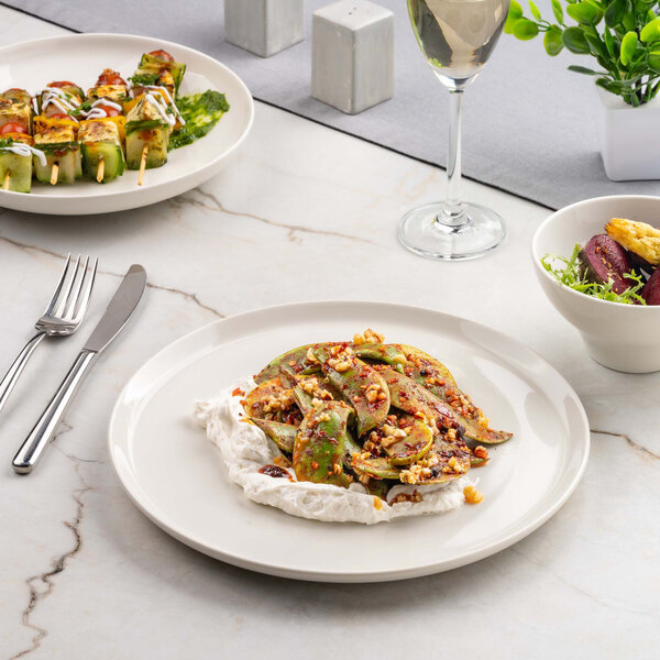 A round ivory porcelain plate with a serving of food, featuring a creamy base topped with sliced vegetables and a nutty garnish.