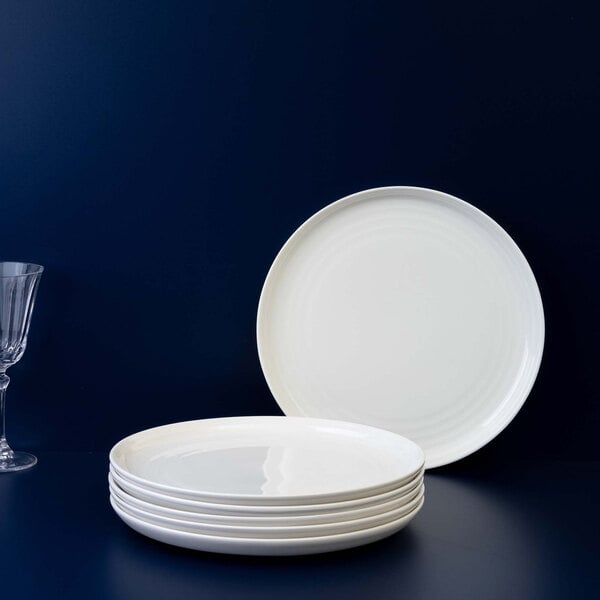 A stack of five round white plates with one plate standing upright behind them, set against a dark blue background with a clear glass on the left side.