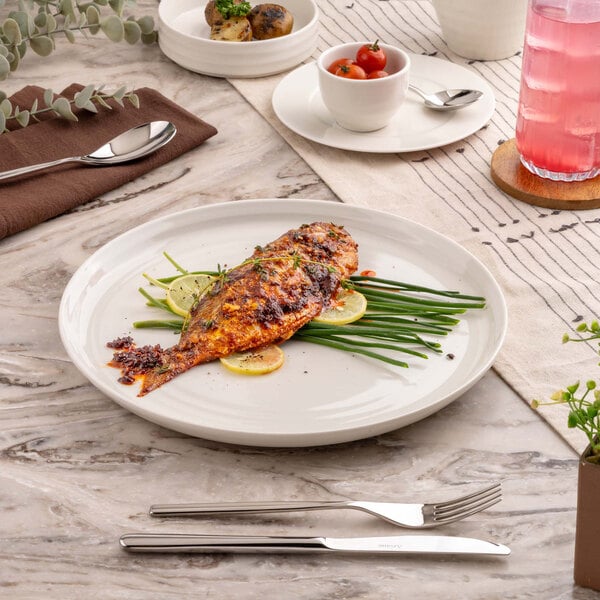 A round white plate with a cooked whole fish garnished with green beans and lemon slices, set on a marble table with cutlery and other dishes nearby.