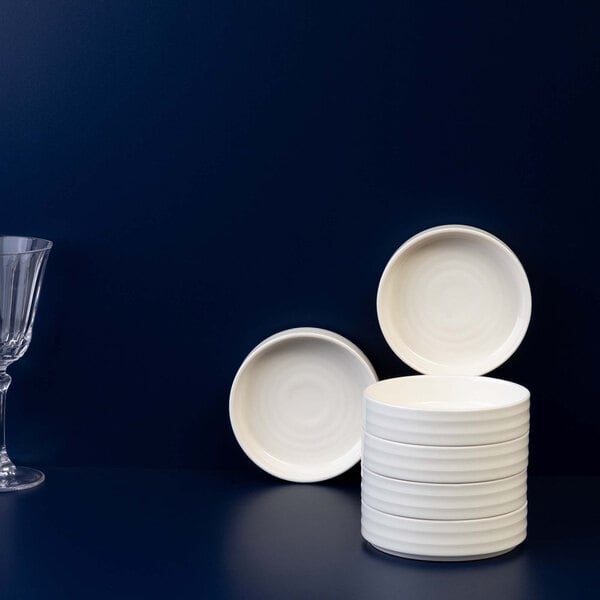 A stack of small, round, white bowls is displayed on a dark blue surface with two additional bowls propped up and a clear glass on the left side.