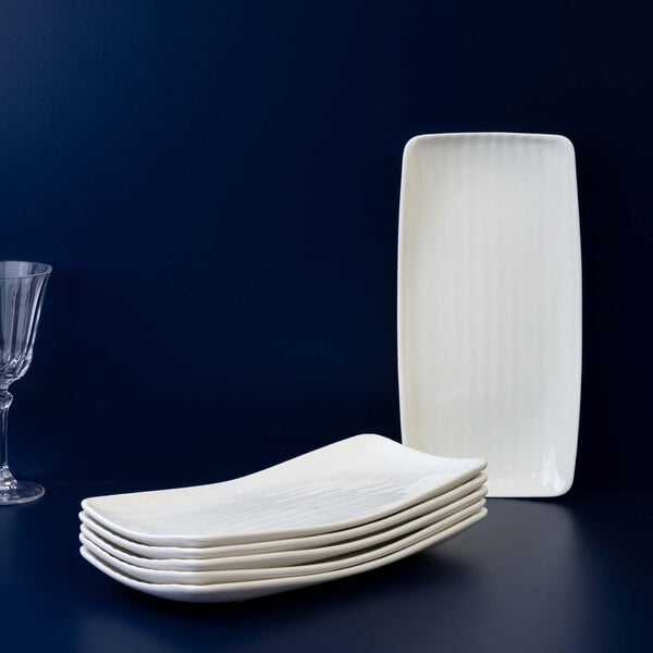 A stack of six rectangular ivory platters with subtle vertical ridges, displayed against a dark background with one platter standing upright and a glass partially visible on the left.