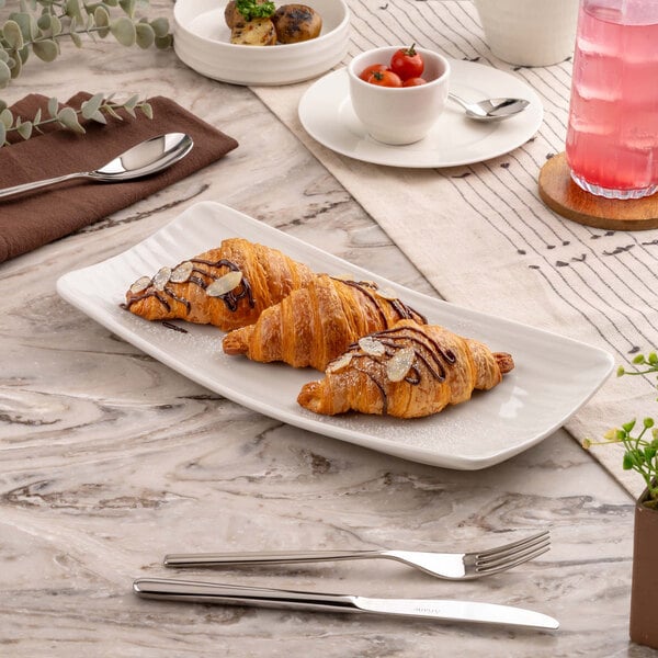 A rectangular white platter with three croissants topped with chocolate drizzle and almond slices, placed on a marble-patterned table with cutlery and other breakfast items nearby.
