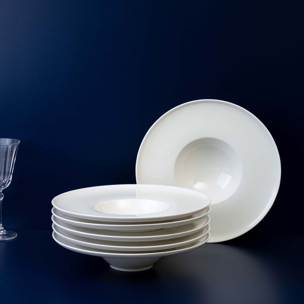 A stack of six white, round, deep plates is displayed next to a single matching plate standing upright, with a glass partially visible on the left side.