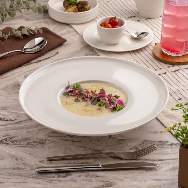 A large white porcelain plate with a small portion of creamy soup garnished with herbs and edible flowers, set on a marble table with cutlery, a pink drink, and other small dishes nearby.