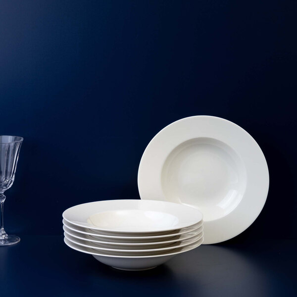A stack of six white deep plates with one plate standing upright behind them, next to a clear wine glass, all set against a dark blue background.