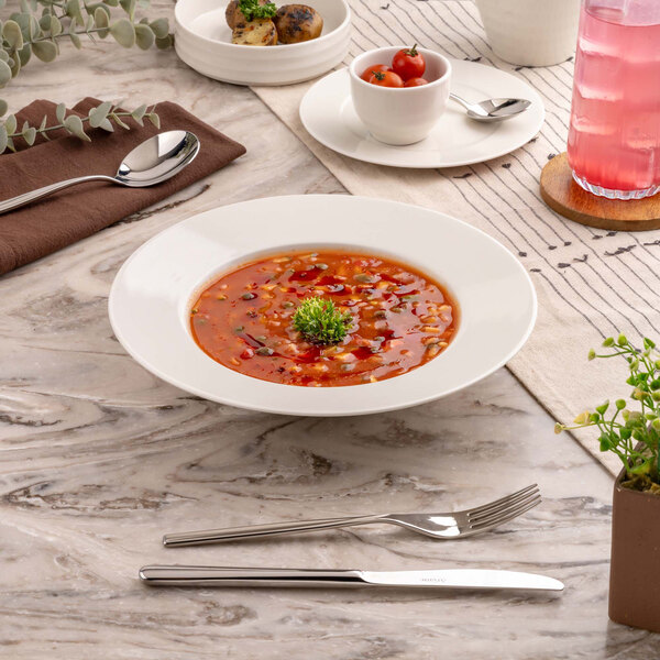 A white, round, deep plate filled with a red soup garnished with herbs, placed on a marble table with cutlery, a napkin, and other tableware nearby.