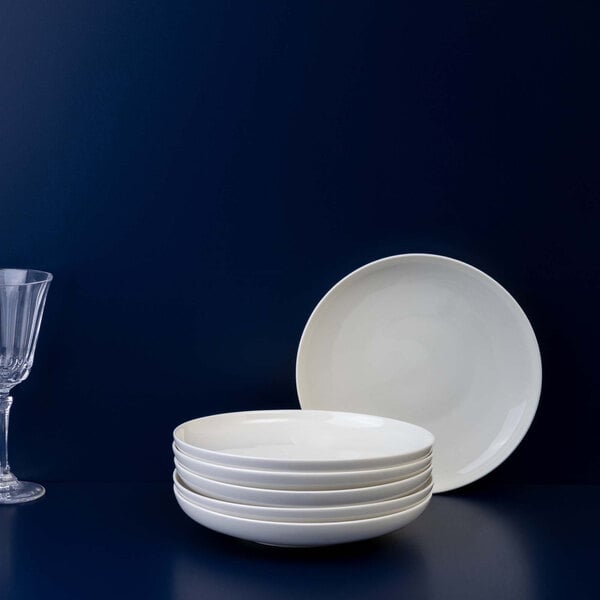 A stack of five white shallow bowls is placed in front of a single upright bowl, with a clear wine glass to the left, all set against a dark blue background.