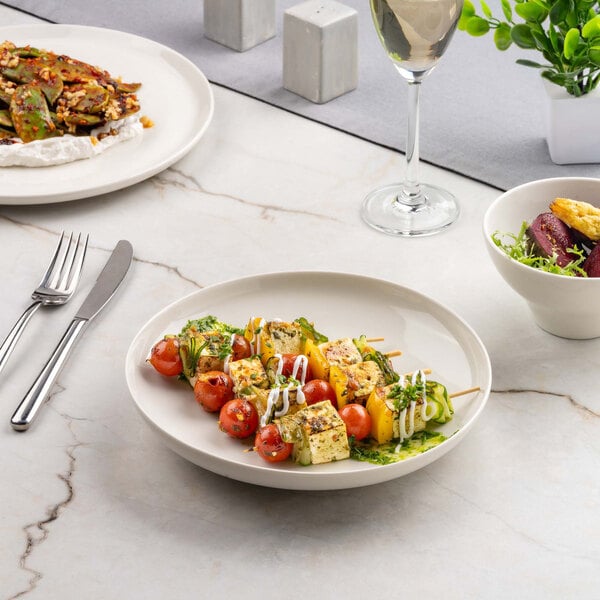 A round ivory porcelain shallow bowl containing skewers of grilled vegetables and paneer, garnished with herbs and sauce, is placed on a marble surface alongside other dishes and a glass of white wine.