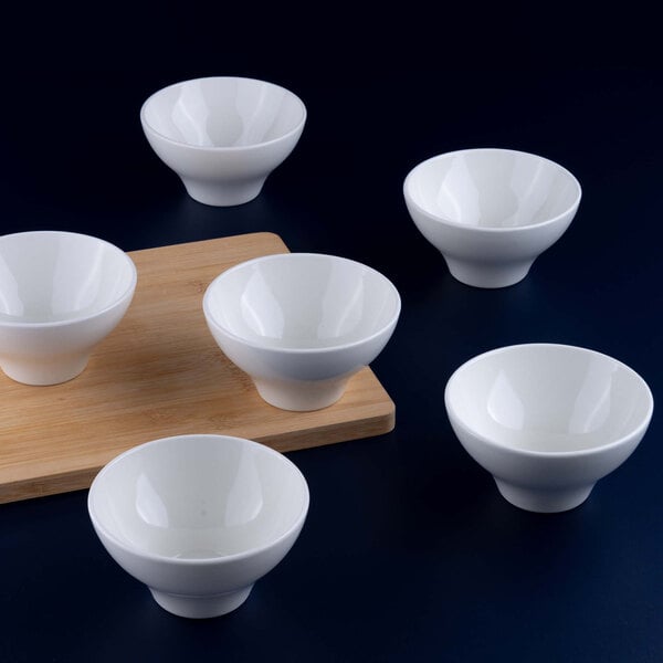 Six small white porcelain bowls are arranged around a wooden board on a dark surface.